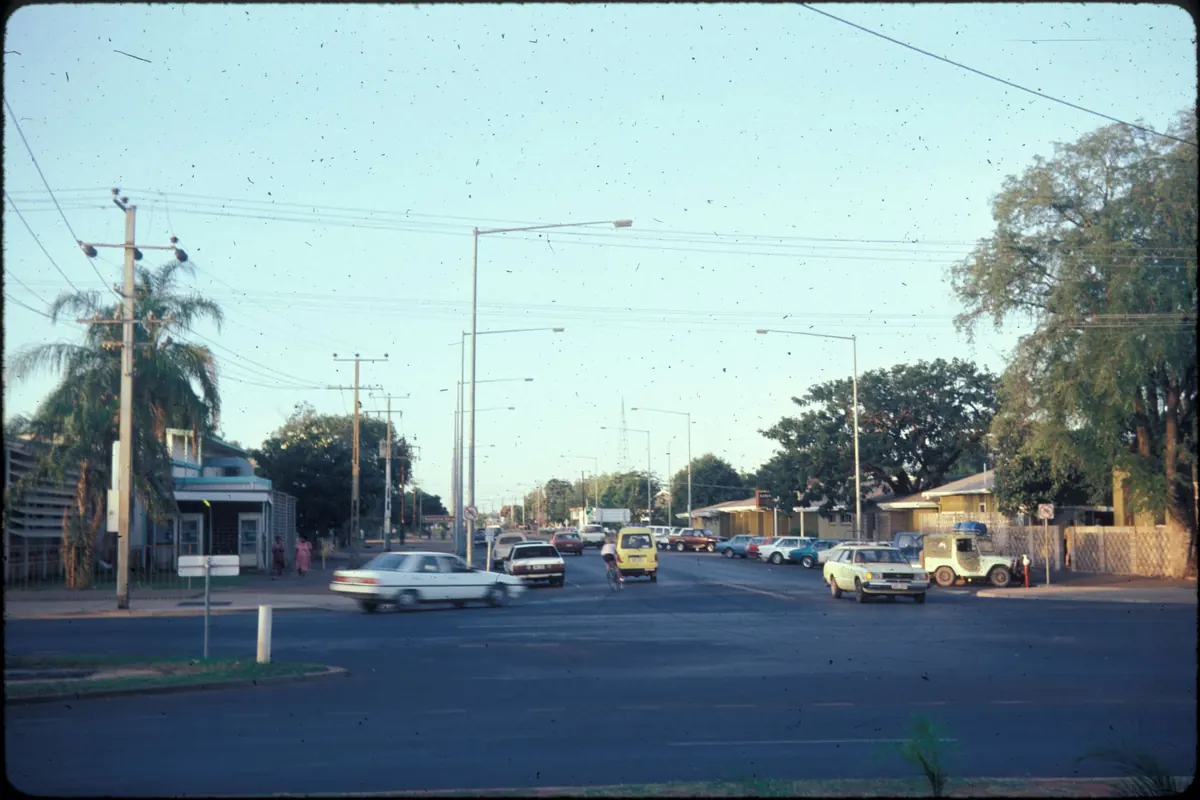 A wide main street with parked cars, power poles, a palm tree and a post office on the left, photographed in 1985